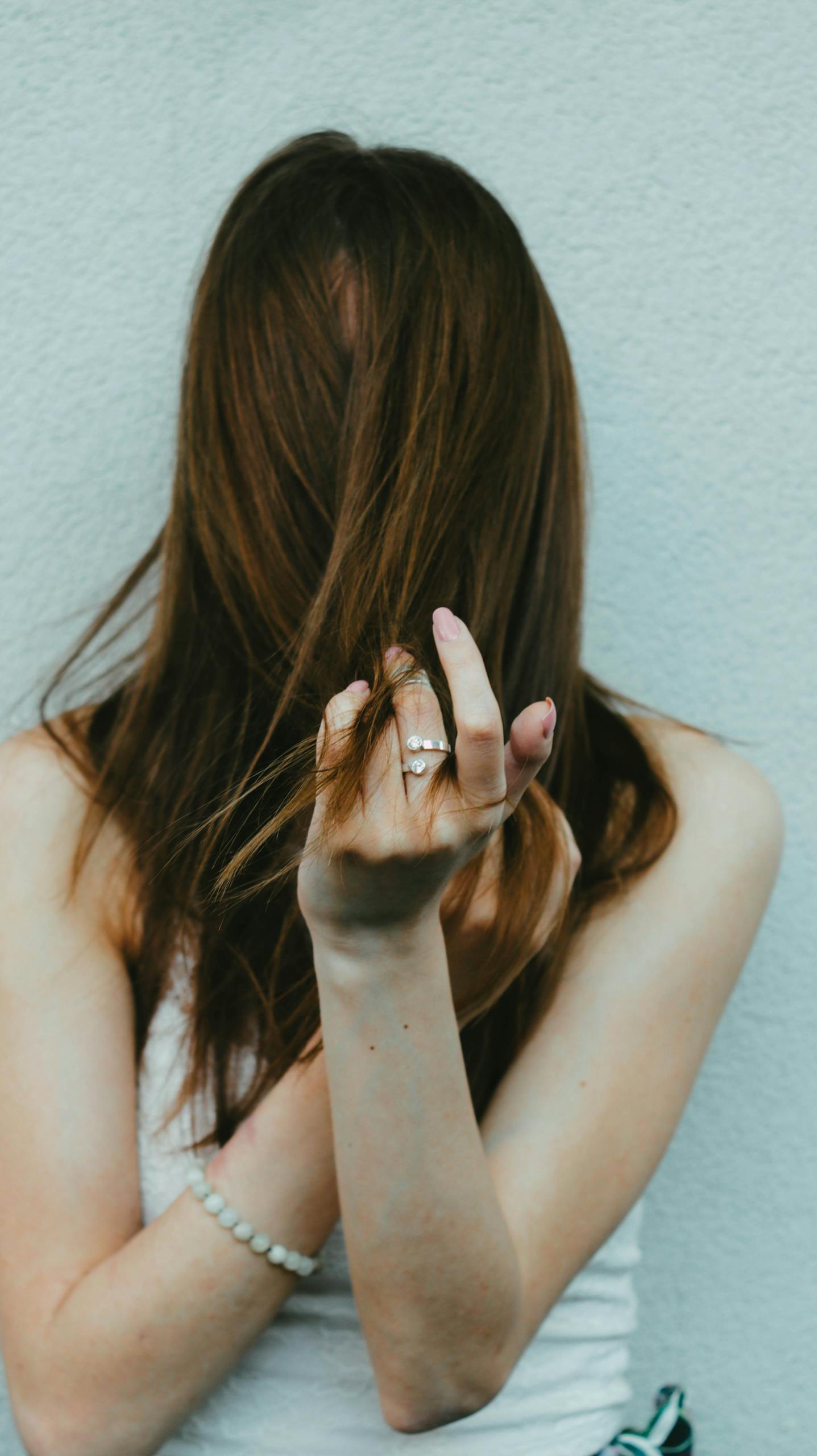 Unrecognizable slim female with hair covering face in ring and wristband standing near cement wall